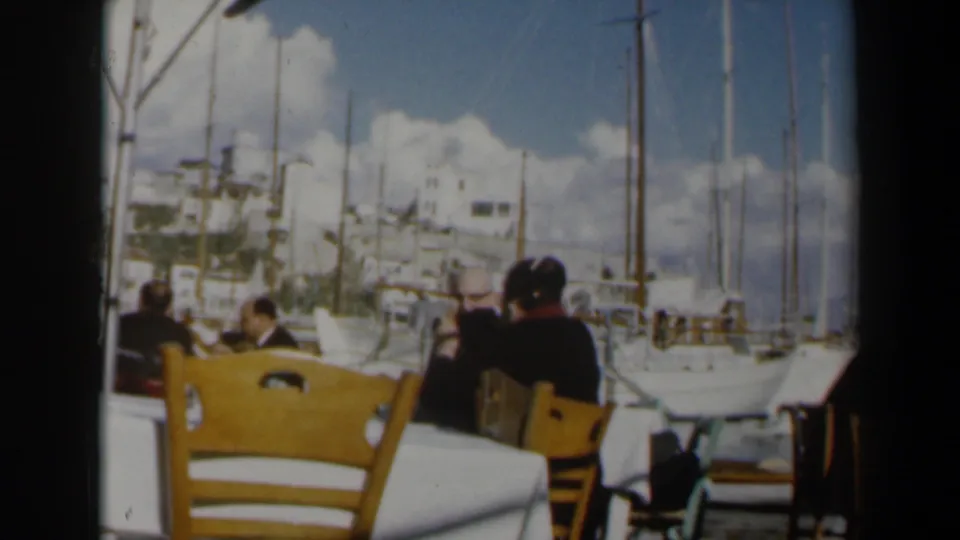 Waterfront restaurant view in Israel, 1960, showing diners from behind with sailboats, masts, sky, and light-colored buildings beyond.
