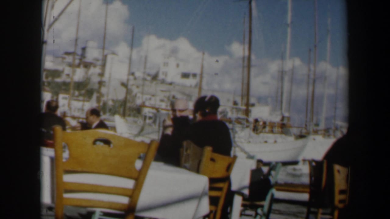 Outdoor restaurant tables beside a marina with seated diners and sailboats in the background.