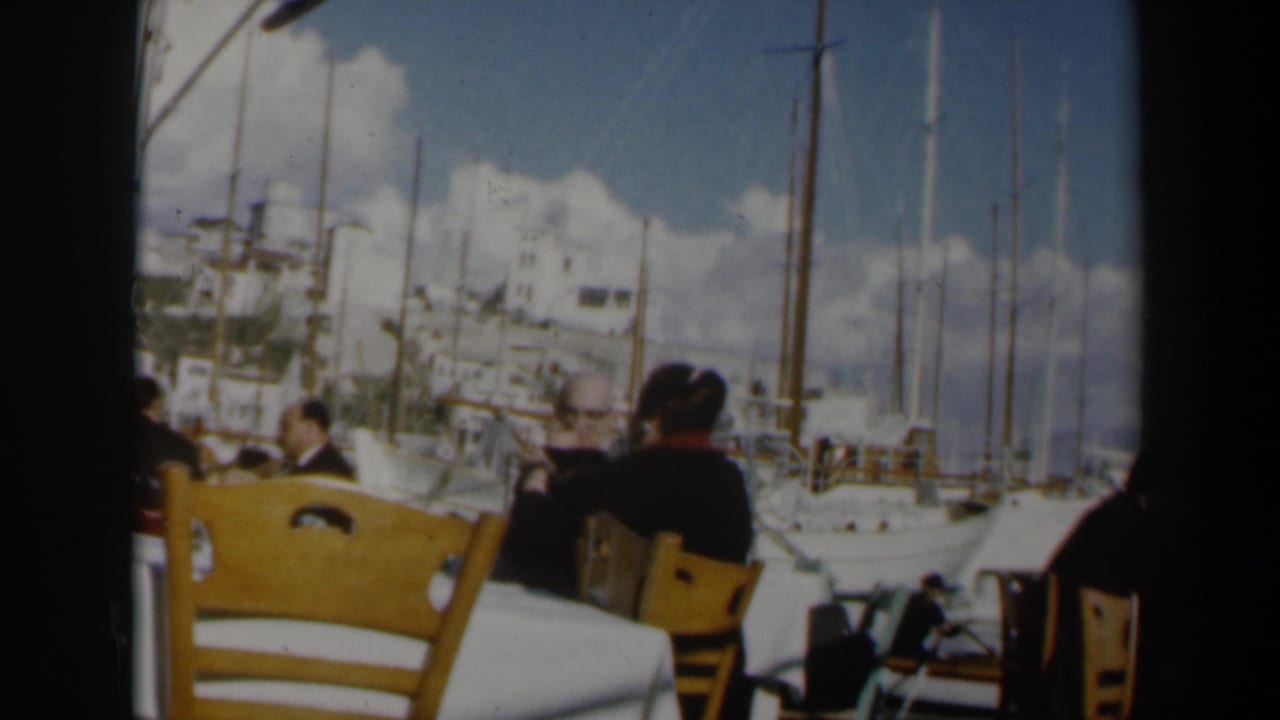 Seated restaurant patrons face a marina with sailboats and white buildings beyond.