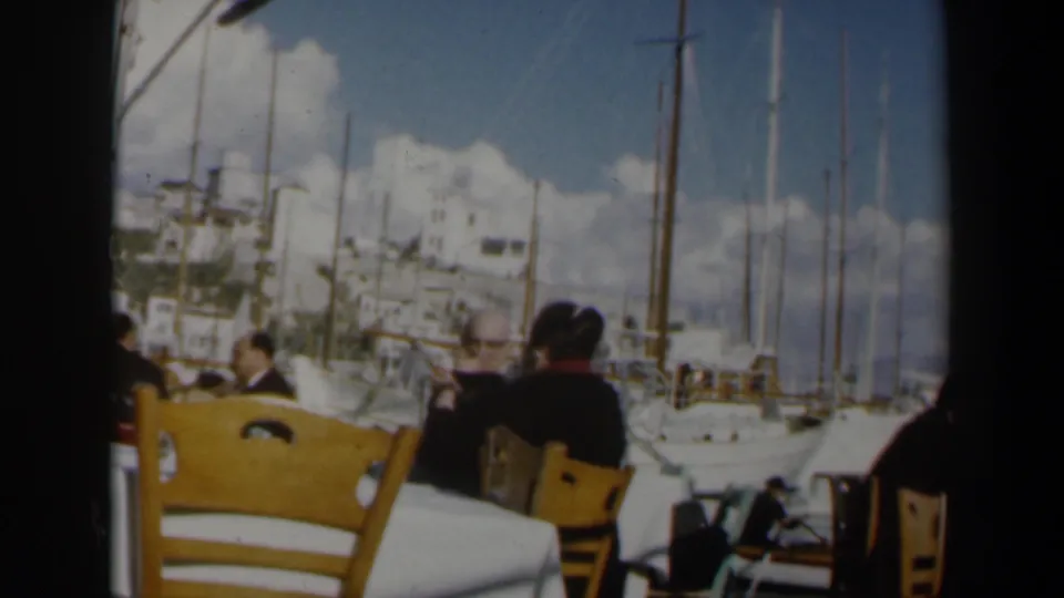 1960 Israel home-movie frame of patrons at an outdoor seaside cafe, with sailboat masts and bright white buildings behind them.