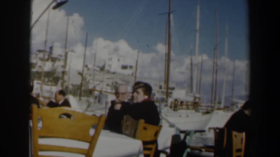 1960 Israel 8mm view of outdoor cafe seating, diners in dark clothing, moored sailboats, tall masts, and bright coastal buildings.