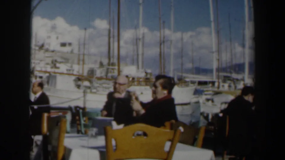 1960 Israel frame showing two visible male patrons at a seaside restaurant, with wooden chairs, a table, sailboats, and marina masts behind them.