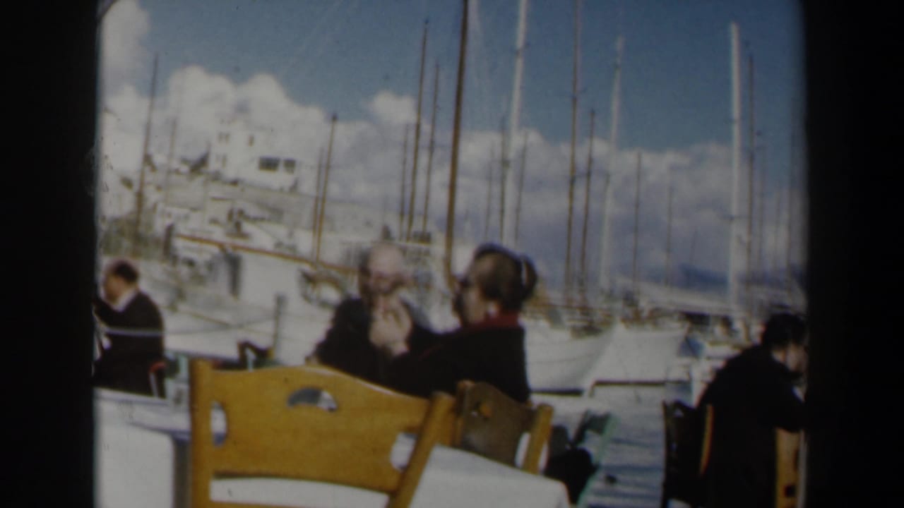 1960 Israel home-movie frame showing adults at an outdoor harbor cafe with wooden chairs, white tablecloths, sailboats, and masts behind them.