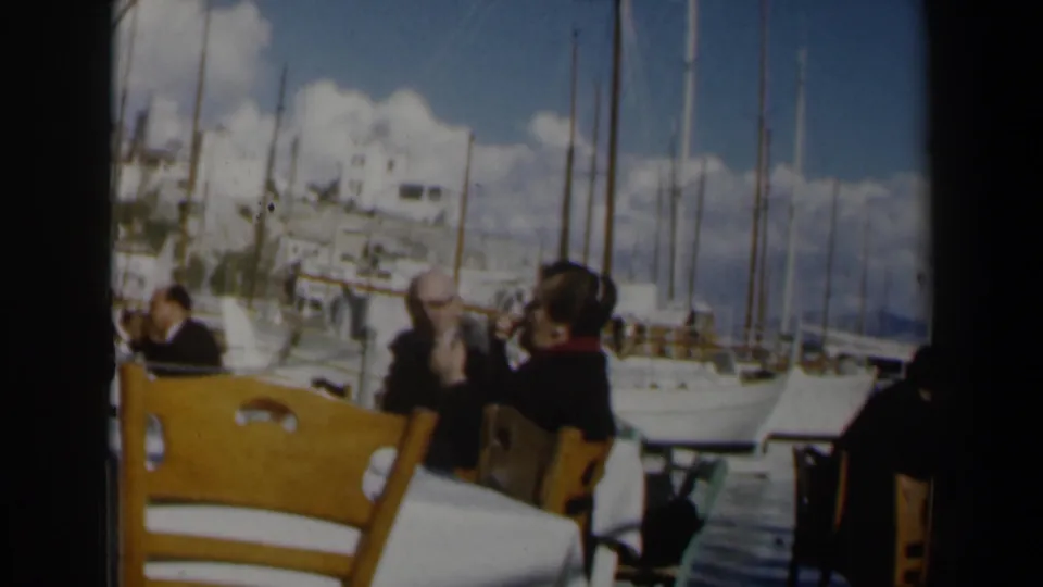 1960 Israel travel footage frame with outdoor cafe seating in the foreground and moored sailboats across the harbor.