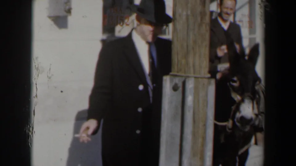 1960 Israel street frame showing a suited man in a dark hat holding a cigarette, with another man and a donkey near a wooden post.