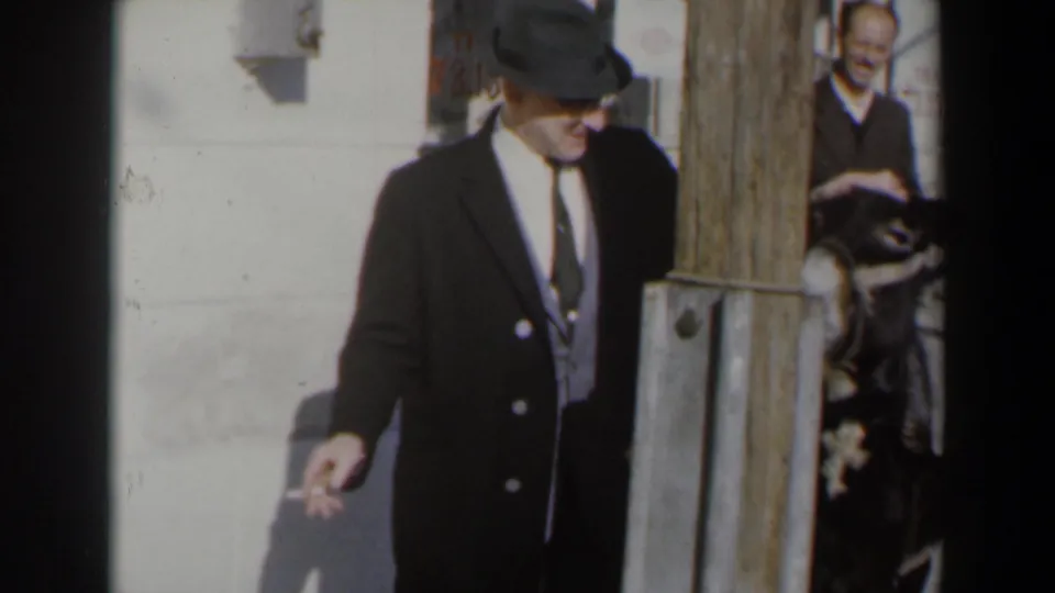 1960 Israel home-movie frame with a man in a dark suit and hat near a wooden post, and a donkey partly visible behind him.