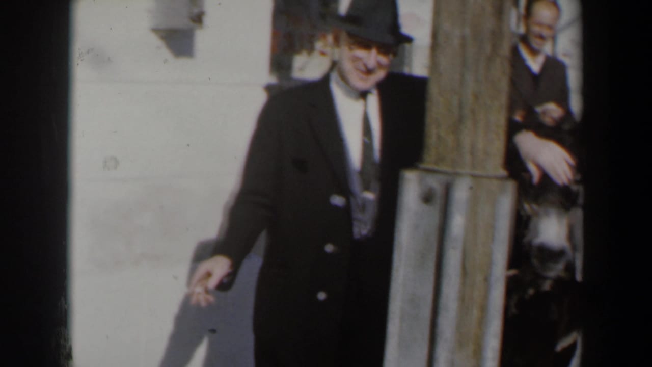 Suited man in a fedora smokes beside a donkey near a wooden post.