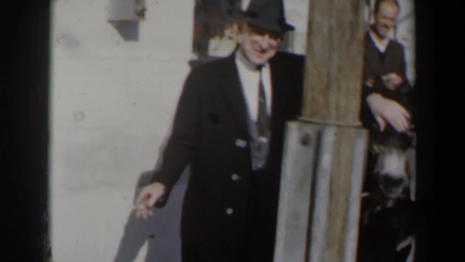 In supported 1960 Israel home-movie footage, a suited man in a fedora smokes while standing beside a donkey and wooden post.