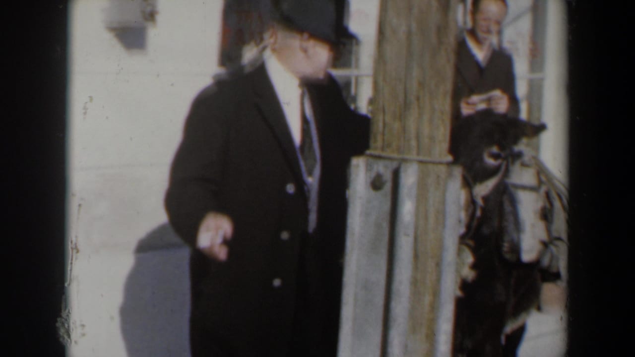 Man in dark suit turns toward a donkey beside a wooden post.