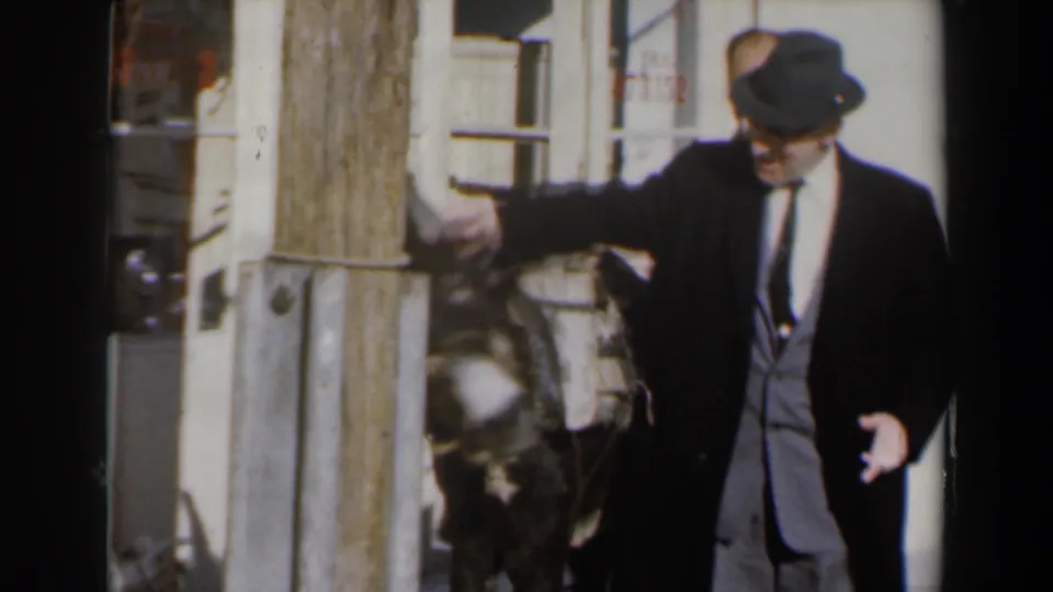 A suited man in a fedora pets or gestures over the donkey near a wooden post, with a small background sign partially visible.