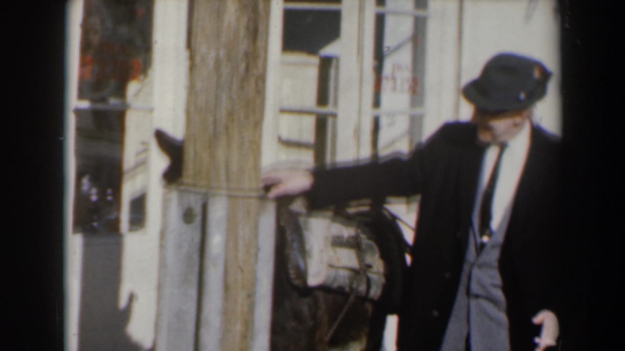 1960s Israel home-movie frame of a suited man in a hat guiding or standing beside a donkey near building facades.