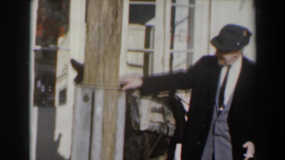 1960s Israel home-movie frame of a suited man in a hat guiding or standing beside a donkey near building facades.