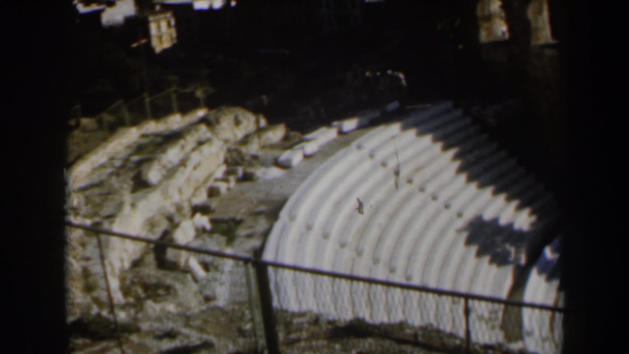 1960 Israel 8mm frame showing curved outdoor amphitheater seating, rough ground, rocks, and a metal railing in strong sunlight.