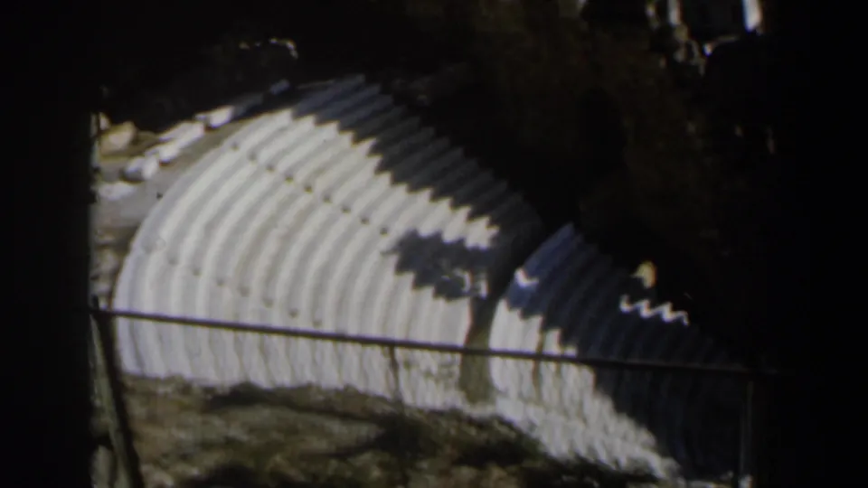 1960 Israel 8mm frame of weathered amphitheater seating, a railing in the foreground, and dark tree or structure shadows across stone rows.