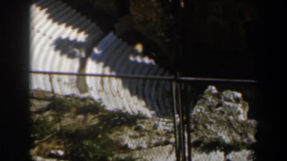 1960 Israel 8mm frame showing part of the amphitheater steps, a foreground rock, metal railings, dry ground, and patches of grass.