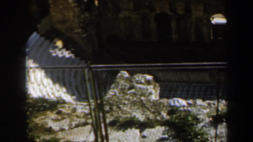 1960 Israel 8mm frame with curved stone seating, foreground railing, rough rocks, dark background masonry, and strong contrast from sunlight.