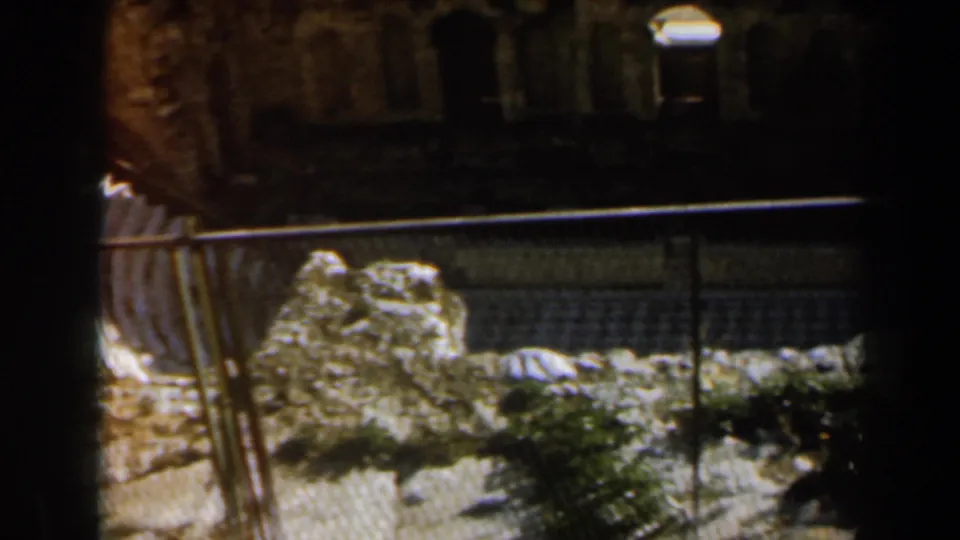 1960 Israel 8mm frame showing an iron fence, sunlit pale stones, low greenery, and dark old stone architecture beyond.