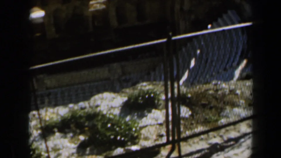 1960 Israel 8mm frame with a curving iron fence, pale ground, low plants, and stone structures beyond.