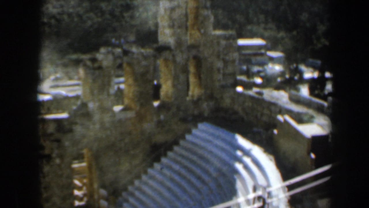 1960 8mm view in Israel looking down over tiered stone amphitheater seating, ruined columns, and a wooded hillside.