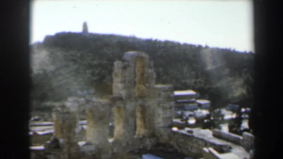 1960 8mm frame from Israel showing weathered stone ruins, a walkway, wooded hillside, and a distant hilltop monument.