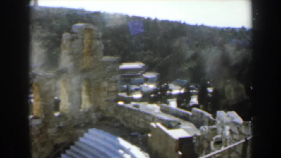 1960 8mm view in Israel across amphitheater seating, broken stone walls, railings, and wooded hills beyond the site.