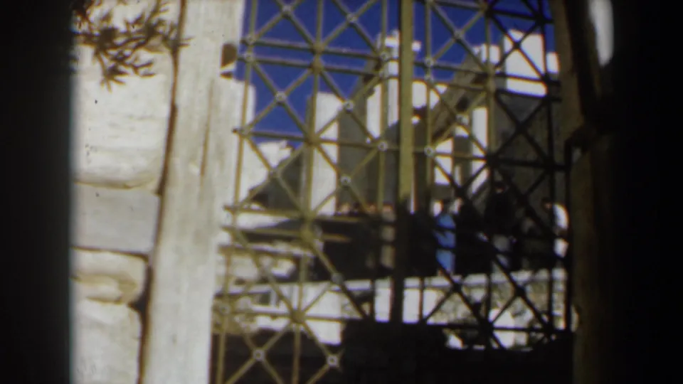1960 8mm frame in Israel showing a metal safety railing or gate beside a pale stone wall, with shadows and partial figures nearby.