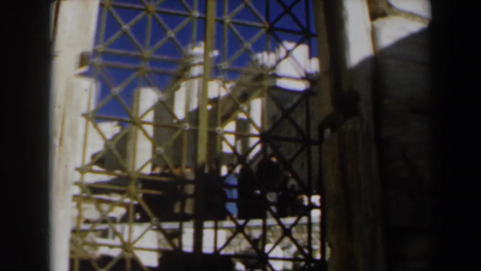 1960s 8mm travel frame showing an iron gate in front of sunlit pale stone architecture and blue sky.
