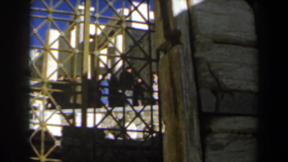 1960s 8mm frame of an iron gate before pale stone ruins, with distant visitors visible in shadow.