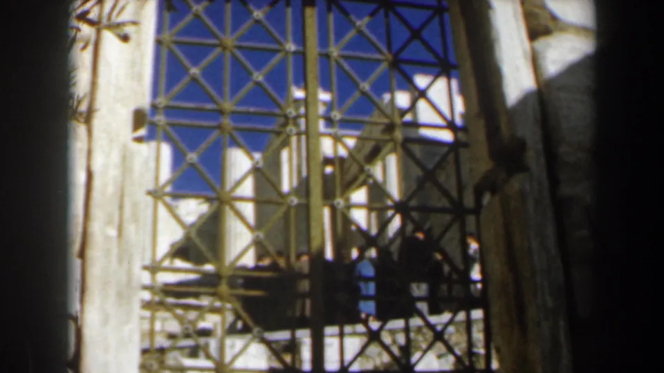 Handheld 1960s 8mm travel view through a patterned iron gate toward sunlit stone architecture.