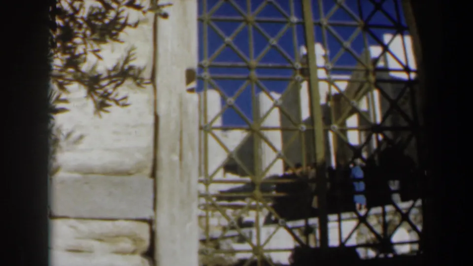 1960s 8mm frame showing a patterned iron gate, pale stone wall, blue sky, and distant visitor figures.