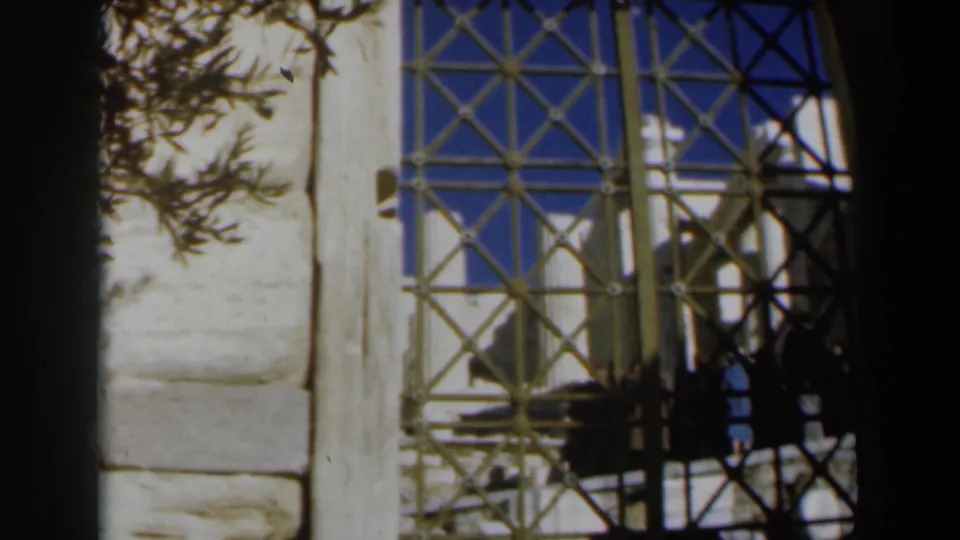1960 Israel 8mm frame looking through decorative tan ironwork, with pale stone columns, blue sky, and dark tree branches at the edge.