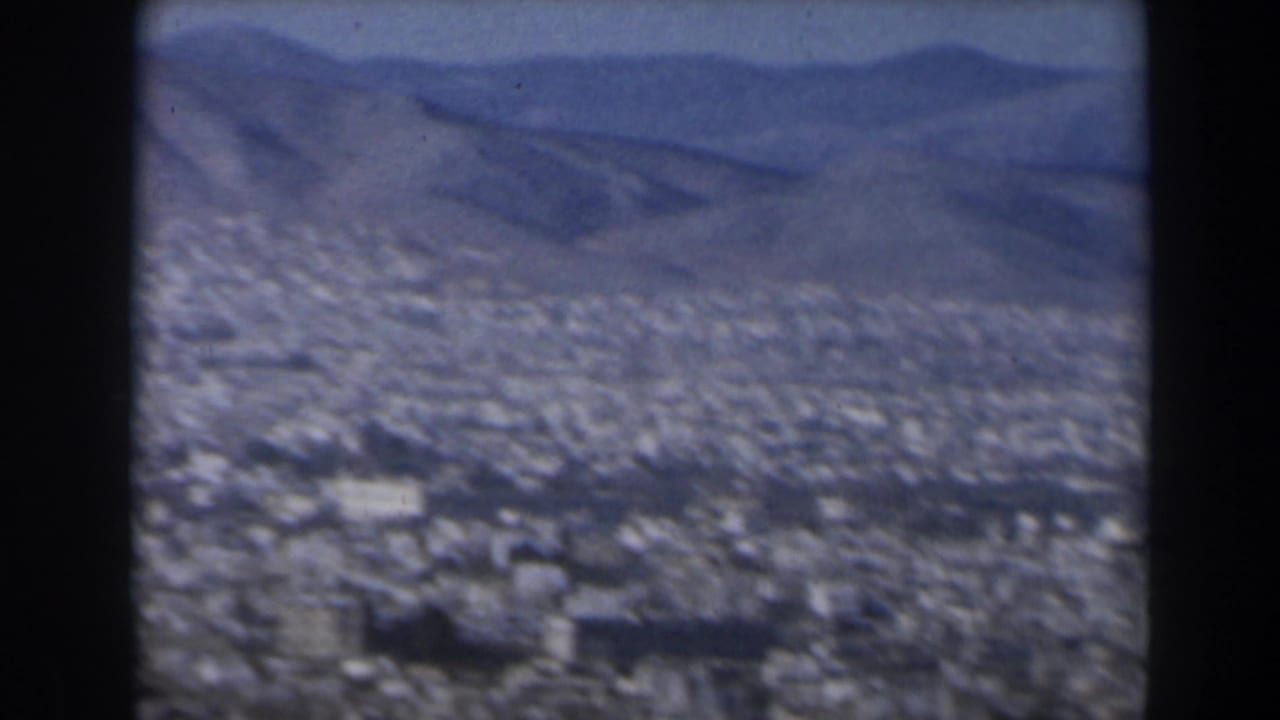 1960 Israel 8mm frame showing a hazy aerial cityscape below distant rugged mountains, seen through dark viewer borders.
