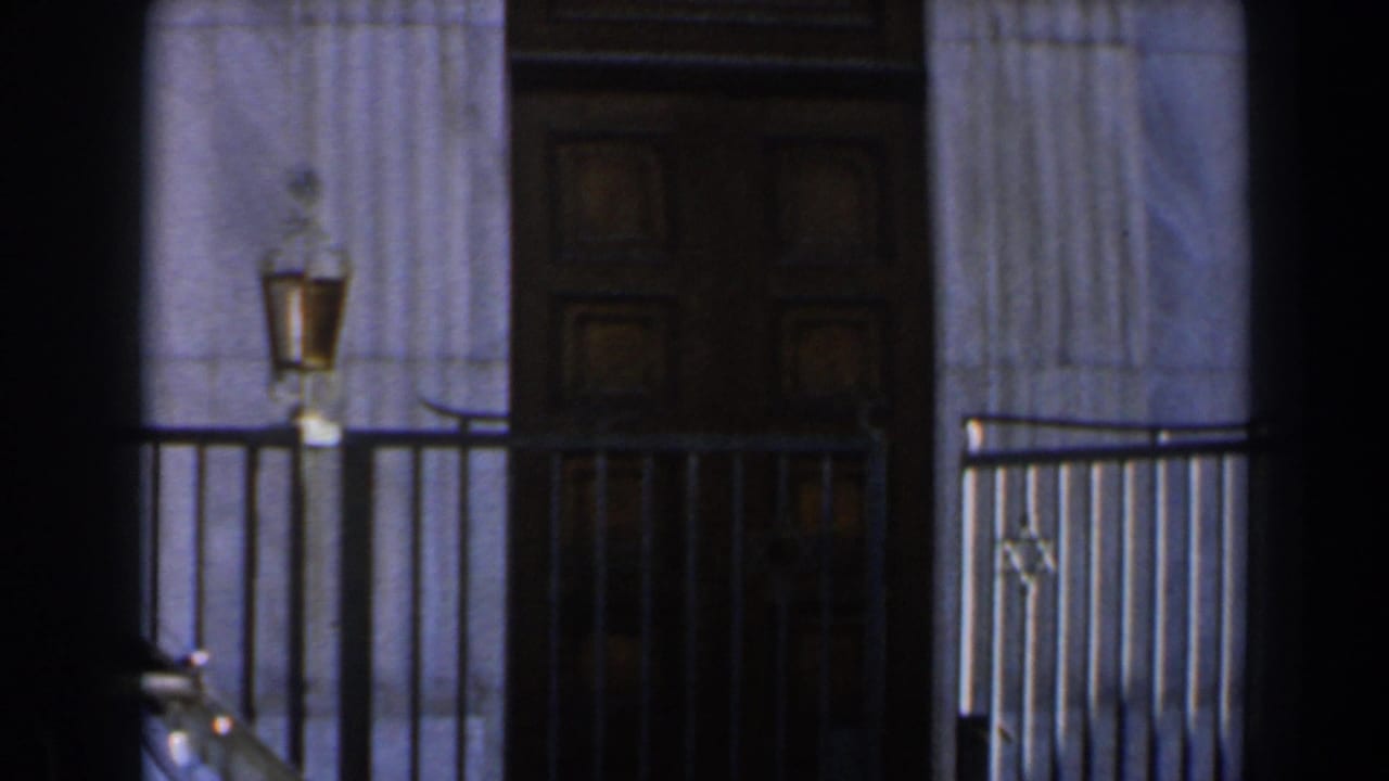 Brown wooden door framed by pale stone columns, metal railings, and wall lanterns.