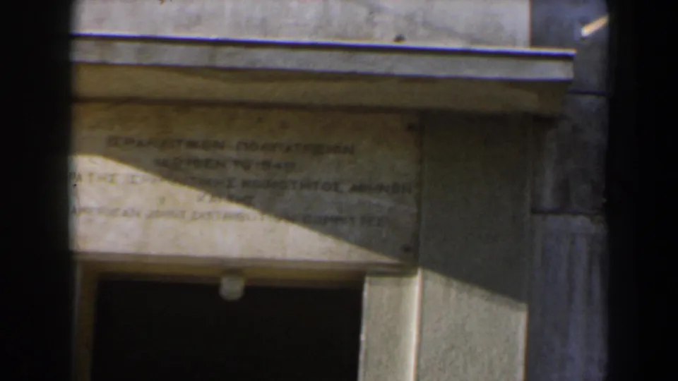 Handheld 1960 Israel frame of the plaque and facade, with a diagonal band of sunlight across the inscription area.