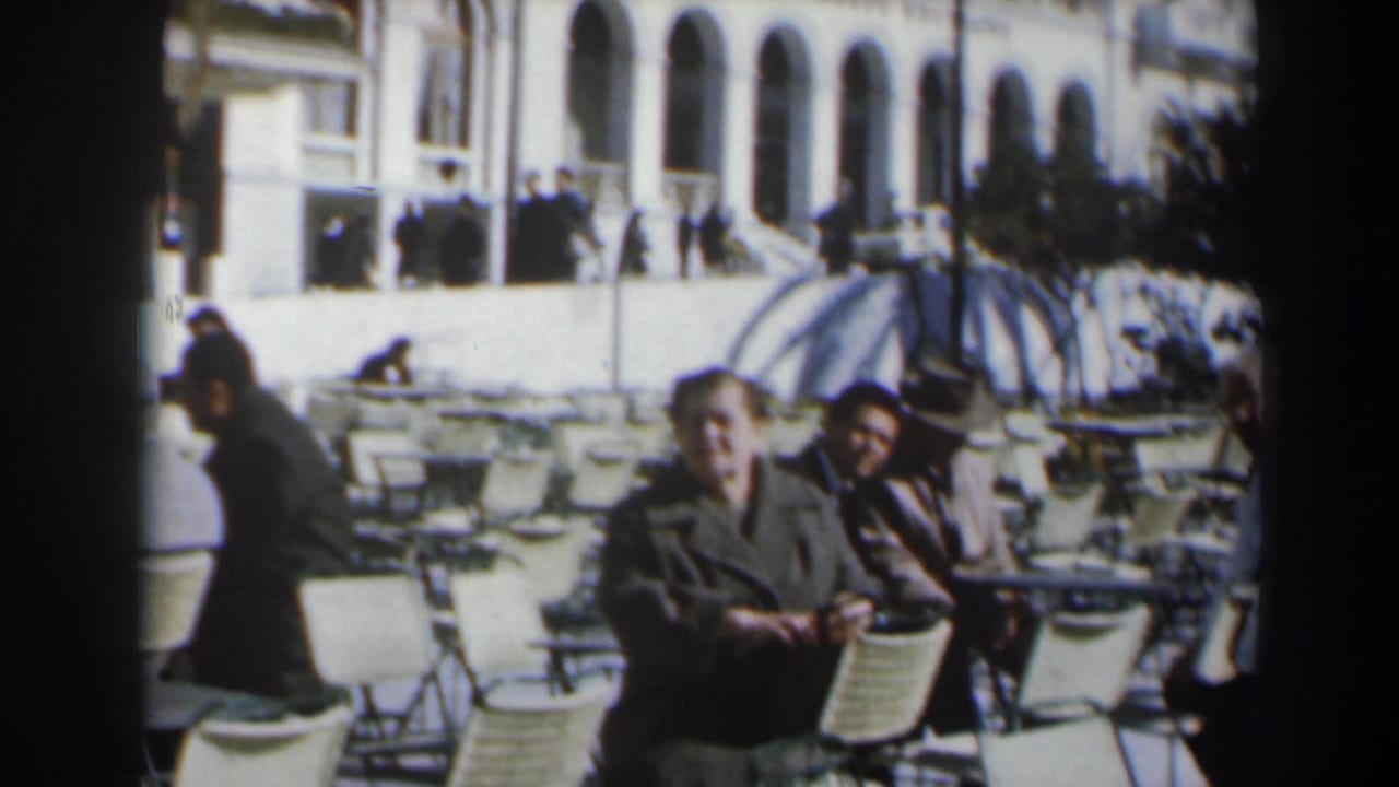 1960s Israel home-movie frame of adults at an outdoor café, with wicker chairs, a parasol, and an arched building in the background.