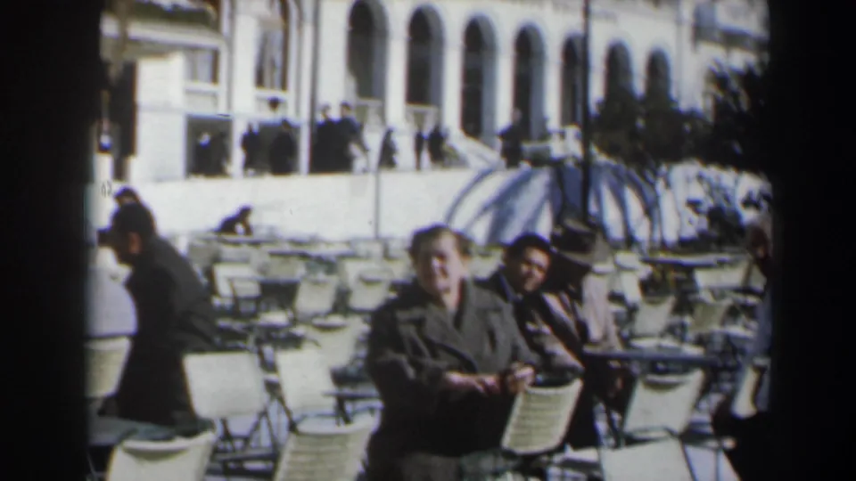 1960s Israel home-movie frame of adults at an outdoor café, with wicker chairs, a parasol, and an arched building in the background.