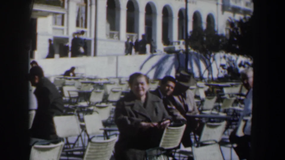 Adults sit among café chairs in a bright outdoor setting in Israel, with a parasol and arched façade visible behind them.