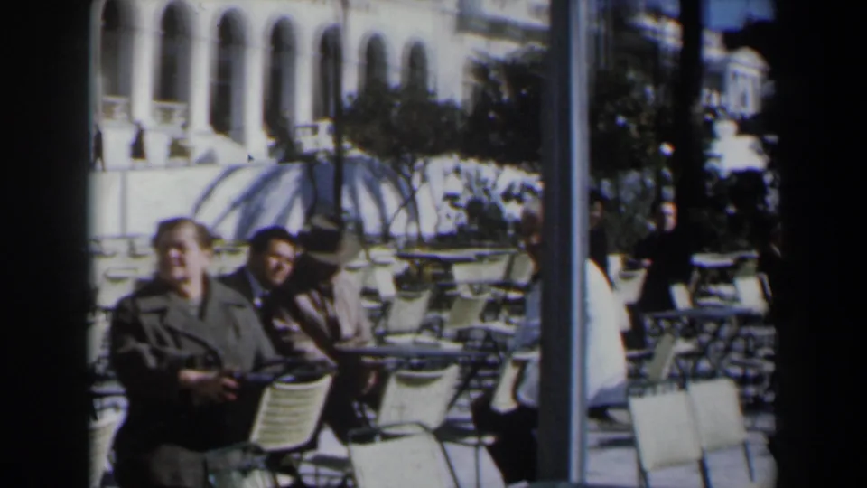A short 8mm frame from Israel shows seated café visitors, many empty chairs, a striped parasol, and an arched building in strong daylight.