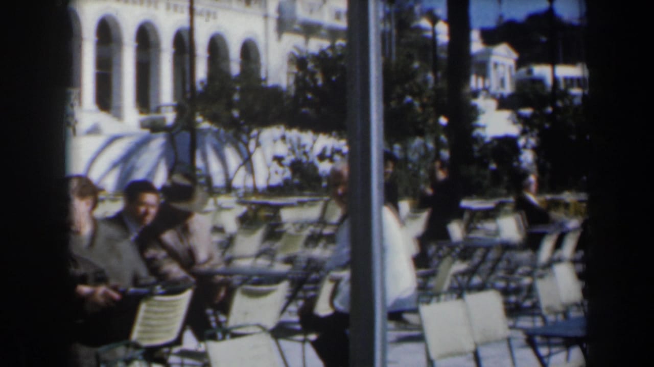 Adults seated at white outdoor cafe tables near an arched building.