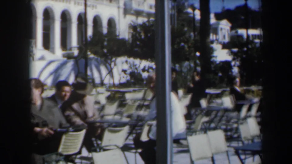 1960s Israel home-movie frame of adults sitting at white metal cafe tables with an arched building facade behind them.