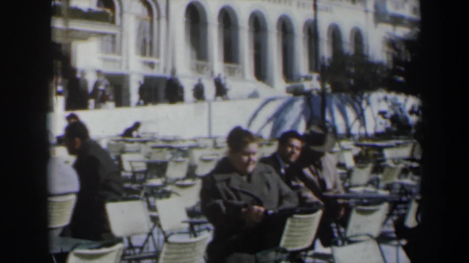 1960s Israel cafe scene with two adults seated in the foreground and rows of white chairs near an arched building.