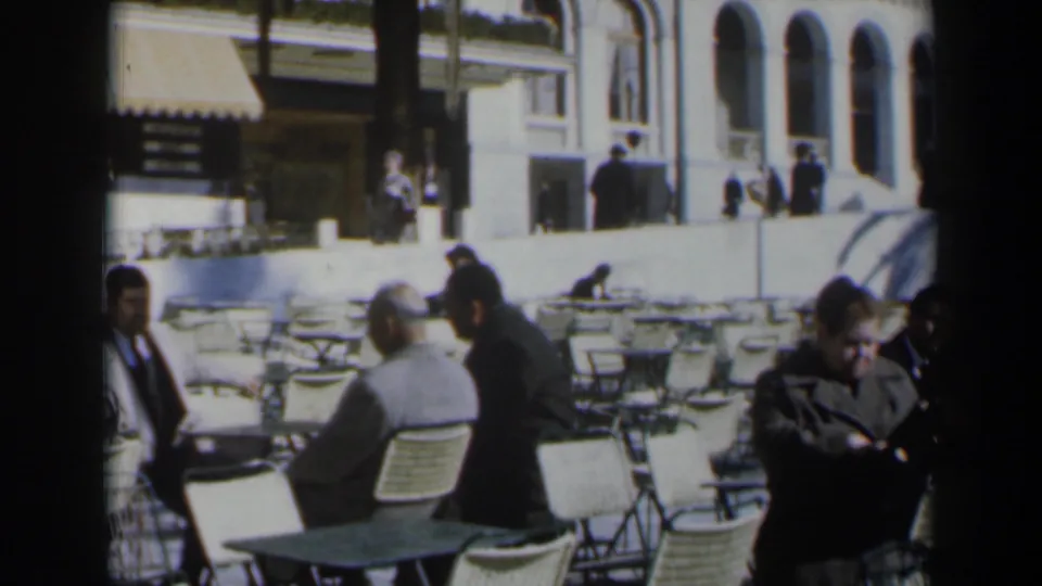 1960s Israel outdoor cafe frame showing seated adults, white chairs, tables, and a standing figure near the edge of the shot.