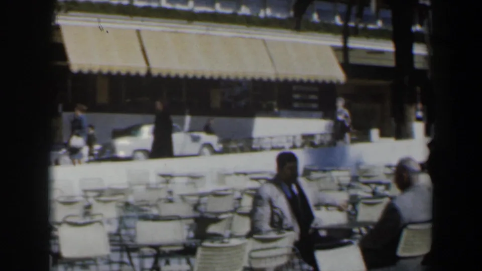 1960s Israel home-movie frame of cafe chairs and patrons with a striped awning and light-colored car behind the seating area.