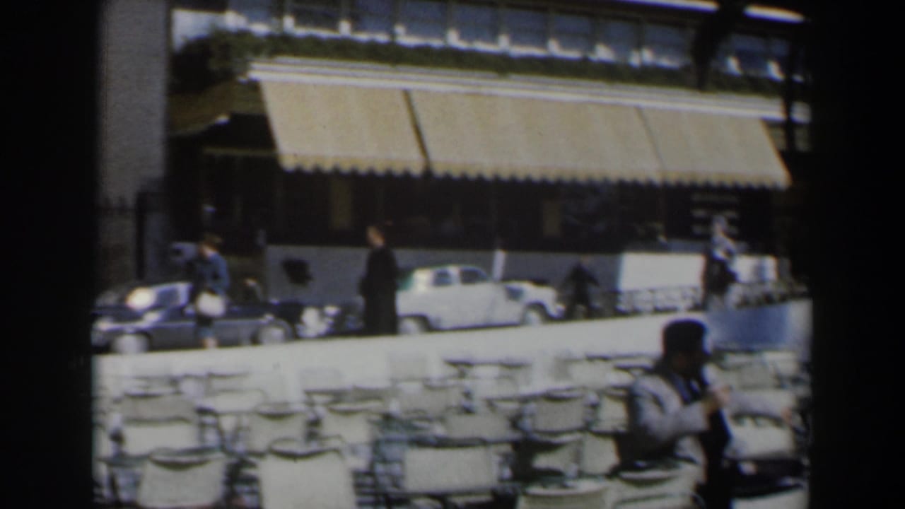 Street view with striped awnings, parked cars, pedestrians, and outdoor chairs.