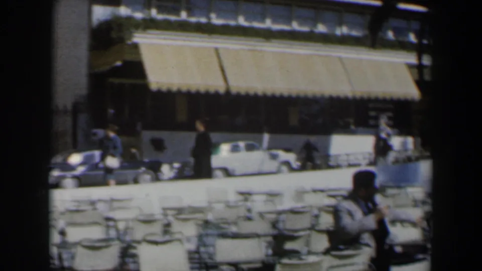 1960 Israel street view with hotel awnings, pedestrians, parked compact cars, and rows of outdoor chairs in the foreground.