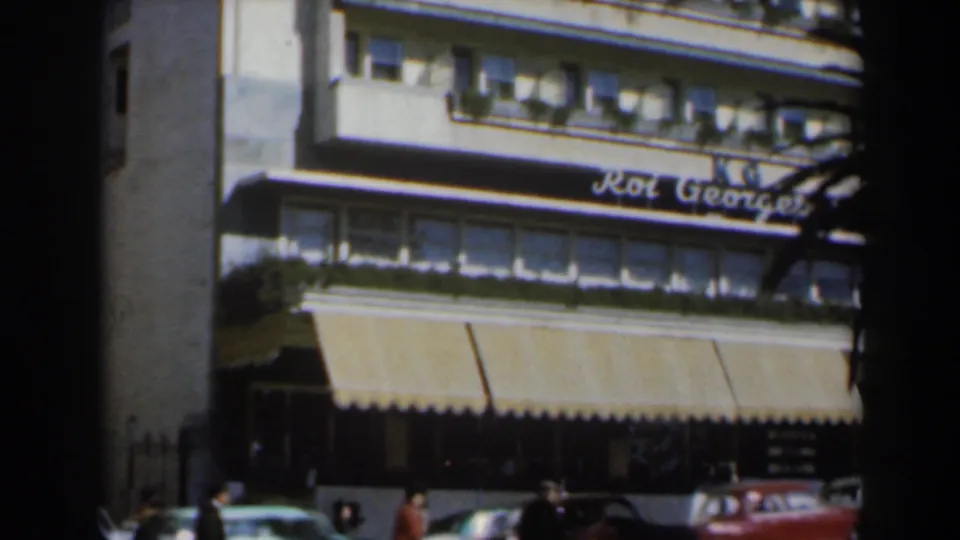 Hotel facade in Israel around 1960 with the Roi George sign, striped awnings, pedestrians, and classic cars along the curb.