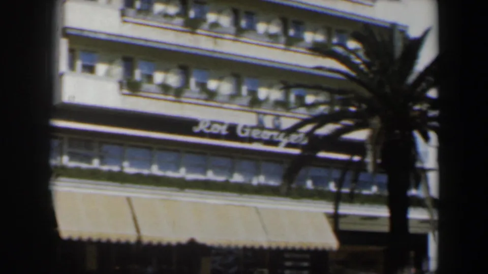Roi George hotel facade with striped awnings and a palm tree crossing the frame, seen in soft 8mm color.