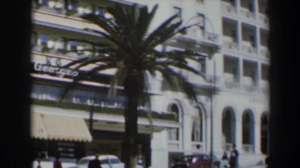 Israel street scene around 1960 with a palm tree, Roi George hotel awnings, neighboring arched facade, pedestrians, and parked cars.