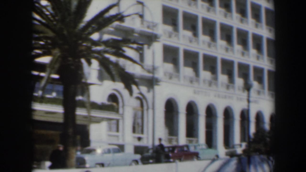 White hotel facade with arches, balconies, palm tree, parked cars, and pedestrians near the street.