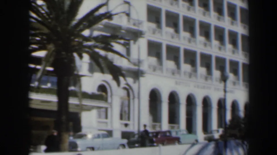 1960 Israel home-movie frame showing Hotel Grand Bretagne facade with arched windows, balconies, palm tree, parked cars, and pedestrians.
